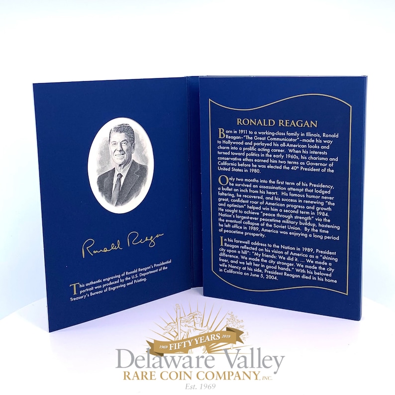 May include: A blue commemorative folder featuring a black and white portrait of Ronald Reagan in an oval frame, his gold signature, and a biography printed in gold text. The folder includes text about his life, presidency, and death in 2004.