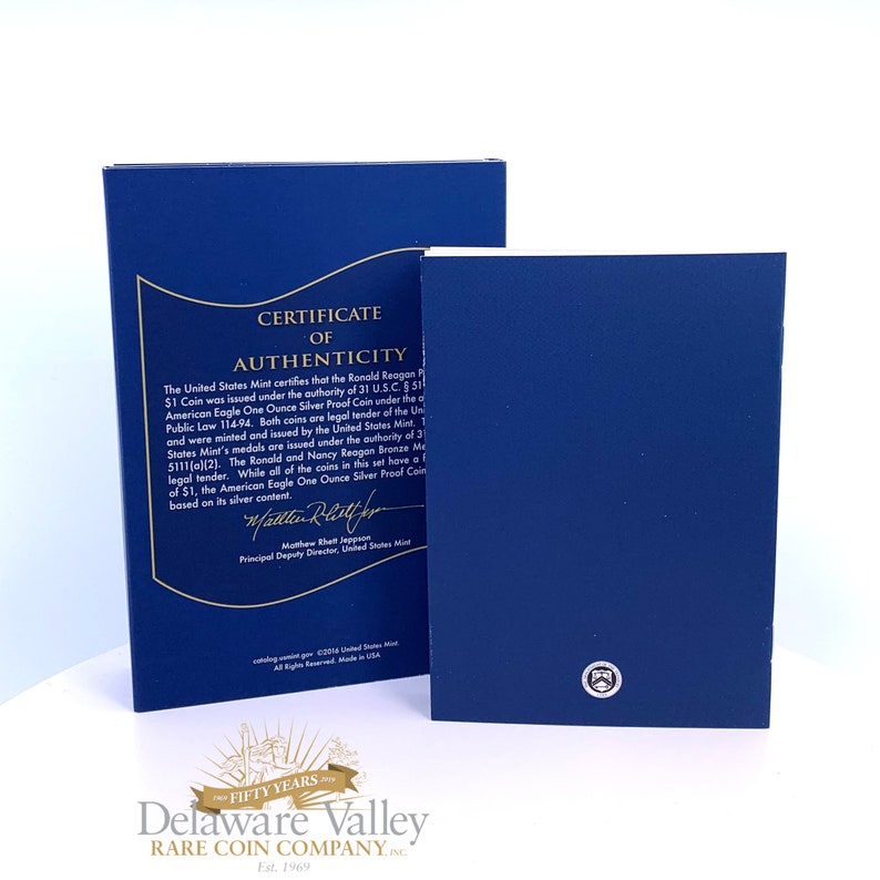 May include: A dark blue United States Mint Certificate of Authenticity folder with gold text and a gold border. The document details the Ronald Reagan $1 Coin and includes a signature from Matthew Rhett Jeppson. A smaller blue folder sits in front.