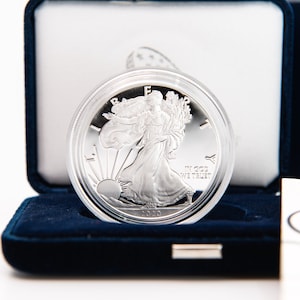 May include: A silver American Eagle coin in a blue velvet box with a white certificate of authenticity. The coin features a depiction of Lady Liberty walking with a torch and olive branch.