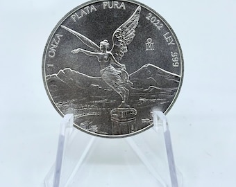 May include: Silver Libertad coin with the Angel of Independence on the front. The coin is on a clear plastic stand. The coin is dated 2022 and says "1 Onza Plata Pura 2022 Ley .999" around the edge.
