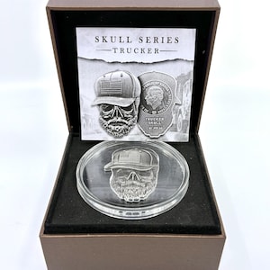 May include: A silver coin featuring a skull wearing a trucker hat with an American flag design, displayed in a brown box. The coin is part of the "Skull Series Trucker" collection, with additional text on the packaging.