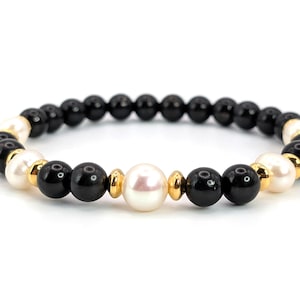 Op de afbeelding: Een elegante armband met zwarte onyx kralen, witte parels en gouden accenten. De armband is geregen op een elastische band, waardoor het een stijlvol accessoire is. De kralen zijn ongeveer 0,8 cm in diameter.
