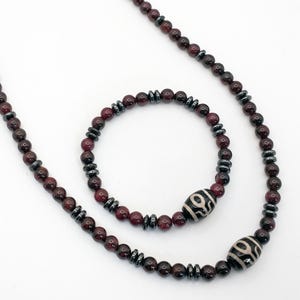 May include: A necklace and bracelet set featuring dark red beads and dark gray accents. The necklace and bracelet also include two white and black patterned beads. The jewelry is displayed on a white background.