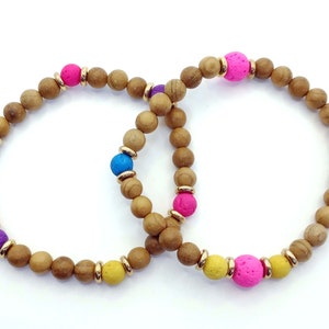 Könnte beinhalten: Zwei Holzperlenarmbänder mit bunten Lavasteinen. Ein Armband hat rosa, gelbe und lila Perlen, während das andere rosa und blaue Perlen hat.