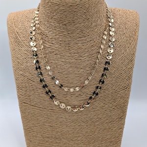 May include: Two gold-colored necklaces displayed on a mannequin. The necklaces are made of small, round, flat, reflective discs. The necklaces are different lengths, with one resting lower than the other. The mannequin is covered in a woven, tan material.