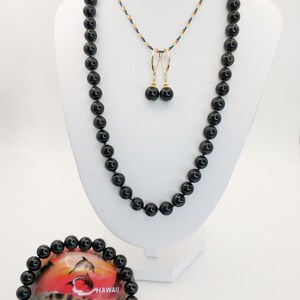Puede incluir: Un conjunto de joyas con un collar de cuentas negras, pendientes y pulsera. El collar y los pendientes se exhiben en un maniquí blanco. La pulsera descansa sobre una concha con un gráfico de delfines y la palabra "HAWAII".