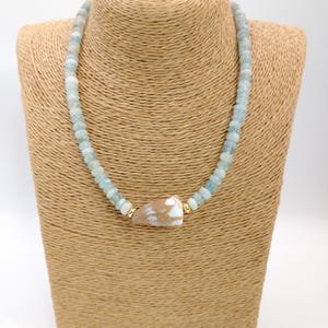 Può includere: Una collana con perline azzurre e un pendente centrale a conchiglia marrone chiaro. La collana è esposta su un busto portagioielli beige. Perline color oro accentuano il design della conchiglia e della collana.