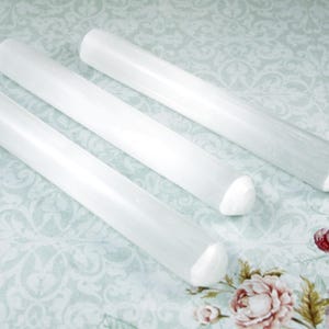 May include: Three white selenite wands with pointed ends. The wands are smooth and polished, and they are lying on a light blue and white patterned surface.