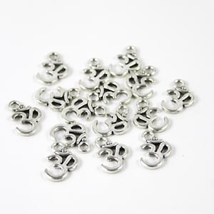 Könnte beinhalten: Silberfarbene Om-Symbol-Anhänger, verstreut auf einer weißen Oberfläche. Die Anhänger haben ein detailliertes Design mit einer Schlaufe oben zum Befestigen an Schmuck oder anderen Bastelarbeiten. Das Om-Symbol ist ein spirituelles Symbol.