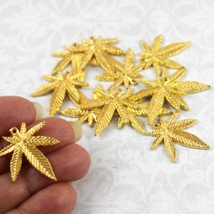 May include: A group of 12 gold-coloured metal cannabis leaf charms. The charms are approximately 2.5 cm in size and have a detailed design. They are ideal for jewellery making, scrapbooking, and other craft projects.