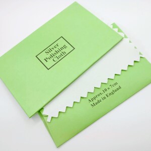 May include: A green envelope with the text "Silver Polishing Cloth" printed on the front. The envelope is open and a white polishing cloth is partially visible. The cloth has a scalloped edge and the text "Approx. 10 x 7cm Made in England" is printed on the envelope.