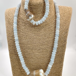 May include: A light blue beaded necklace and matching bracelet set. The necklace and bracelet feature small, round, light blue beads and are accented with a large, cone-shaped, brown and white shell. The jewelry is displayed on a tan bust.