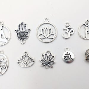 Puede incluir: Surtido de dijes de metal plateado con varios símbolos y diseños, incluyendo una flor de loto, el símbolo Om, una figura meditando y una mano de Fátima.