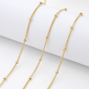 Op de afbeelding: Close-up van verschillende strengen delicate goudkleurige ketting. Elke ketting heeft een fijn curb-link ontwerp afgewisseld met kleine, gelijkmatig verdeelde gouden kralen. De kettingen zijn diagonaal gerangschikt tegen een witte achtergrond, wat een gevoel van elegantie creëert.