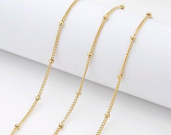 May include: Close-up of several strands of delicate gold-colored chain. Each chain features a fine curb link design interspersed with small, evenly spaced gold beads. The chains are arranged diagonally against a white background, creating a sense of elegance.