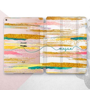 Puede incluir: Una funda blanca para iPad con un diseño abstracto dorado y rosa. La funda tiene una solapa que se pliega con el nombre "Megan" escrito en cursiva en la parte delantera.