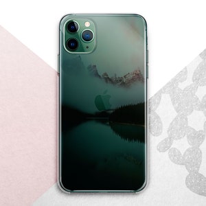 Pu&ograve; includere: Una custodia per telefono trasparente con una scena di montagna verde e blu e un riflesso di lago. La custodia ha un logo Apple nero e un rigonfiamento della fotocamera nero con tre obiettivi.
