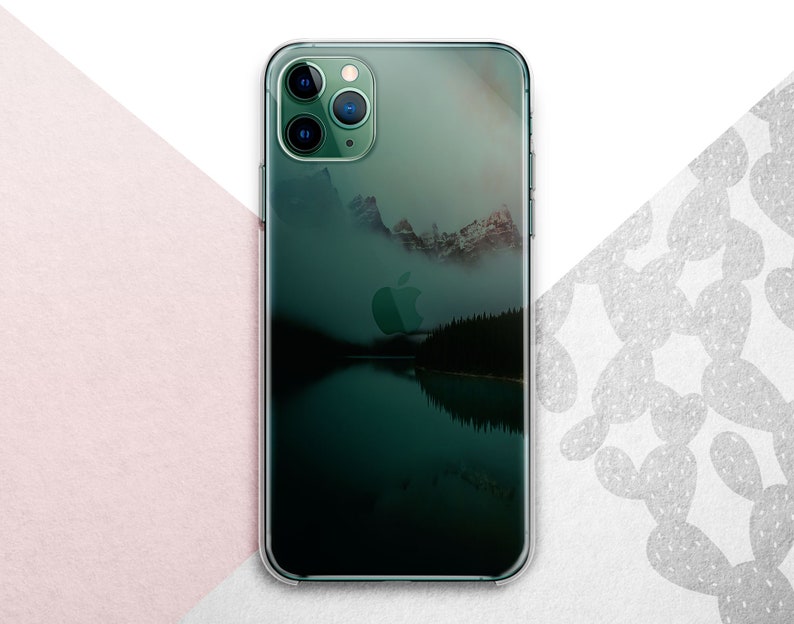 Pu&ograve; includere: Una custodia per telefono trasparente con una scena di montagna verde e blu e un riflesso di lago. La custodia ha un logo Apple nero e un rigonfiamento della fotocamera nero con tre obiettivi.