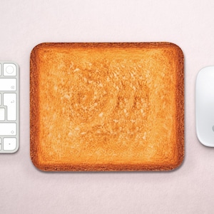 May include: A rectangular mouse pad featuring a realistic, high-resolution print of a slice of golden-brown toasted bread with a textured, porous surface and darker, crispy edges, positioned between a white computer keyboard and a white mouse.