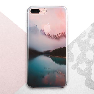 Pu&ograve; includere: Una custodia per telefono trasparente con un'immagine panoramica di una catena montuosa riflessa in un lago. Le montagne sono coperte di nebbia e il cielo &egrave; di un rosa e un arancio tenue.
