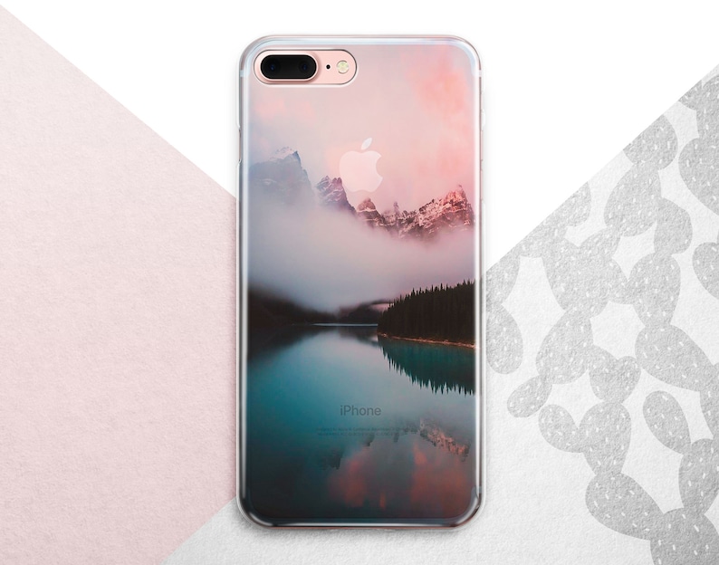 Pu&ograve; includere: Una custodia per telefono trasparente con un'immagine panoramica di una catena montuosa riflessa in un lago. Le montagne sono coperte di nebbia e il cielo &egrave; di un rosa e un arancio tenue.