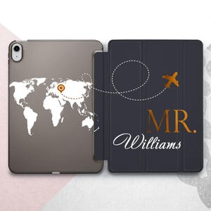 Puede incluir: Una funda gris para iPad con un diseño de mapa del mundo en blanco y un marcador de ubicación dorado. La cubierta presenta el texto "MR. Williams" en dorado y blanco, con una línea punteada y un avión.