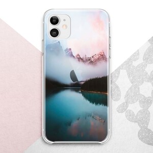 Pu&ograve; includere: Una custodia per telefono trasparente con un'immagine panoramica di un lago di montagna e montagne innevate sullo sfondo. Il cielo &egrave; di un rosa e un arancio tenue.