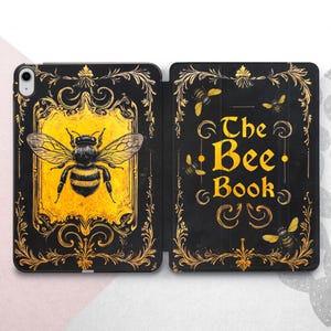 Pode incluir: Capa para iPad preta e dourada com uma ilustração de abelha e o texto "The Bee Book". A capa tem um design vintage com detalhes florais dourados e um fundo amarelo. O design está em ambos os painéis.
