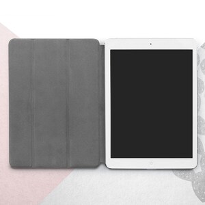 Peut inclure: Une tablette blanche avec un &eacute;cran noir est ouverte, &agrave; c&ocirc;t&eacute; d'une housse de tablette grise. La housse est repli&eacute;e, r&eacute;v&eacute;lant l'&eacute;cran de la tablette. Le fond est un m&eacute;lange de blanc, de rose et de gris.