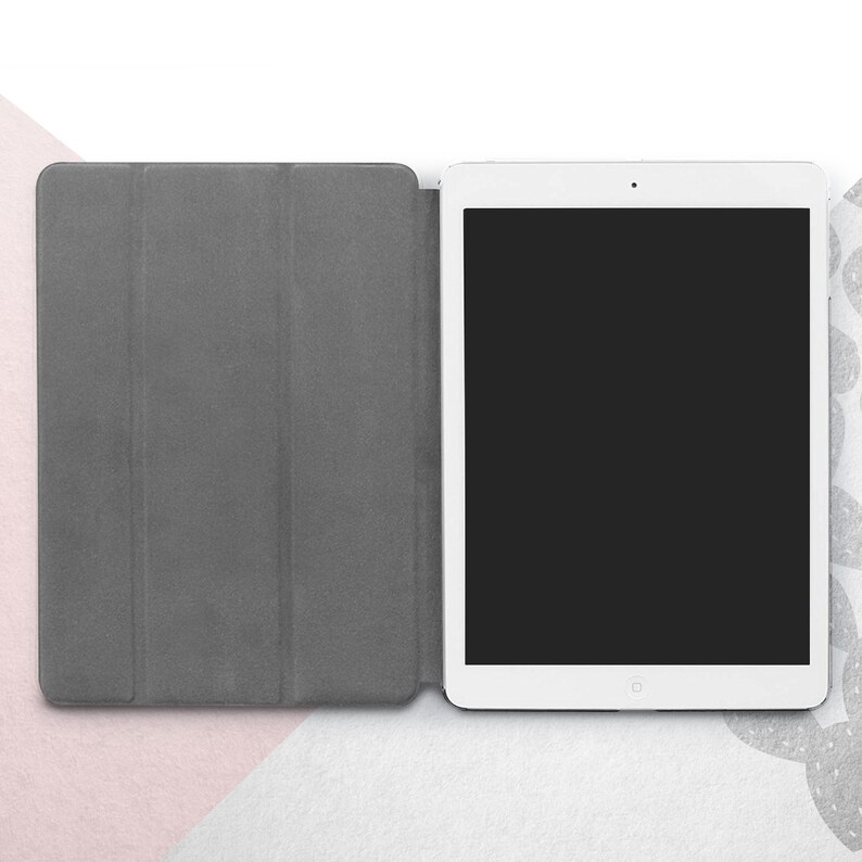 Peut inclure: Une tablette blanche avec un &eacute;cran noir est ouverte, &agrave; c&ocirc;t&eacute; d'une housse de tablette grise. La housse est repli&eacute;e, r&eacute;v&eacute;lant l'&eacute;cran de la tablette. Le fond est un m&eacute;lange de blanc, de rose et de gris.