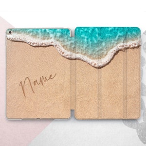 Op de afbeelding: Een gepersonaliseerde iPad-hoes met een strandscene. De hoes toont een close-up van een zandstrand met turquoise water en witte schuimende golven. Het woord "Name" is in een cursief schrift op het zand geschreven.