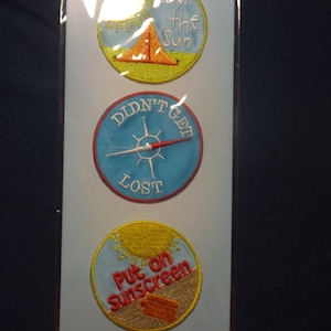 May include: Three embroidered patches with travel-themed designs. One patch says "Soak the Sun" with a tent graphic. Another says "Didn't Get Lost" with a compass. The last says "Put on Sunscreen" with a sunscreen bottle.