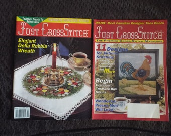 May include: Two copies of "Just CrossStitch" magazine are displayed. One features a Della Robbia wreath, a candle, and a teacup. The other showcases a rooster cross-stitch design, with text that reads "11 Designs for Autumn Stitching."