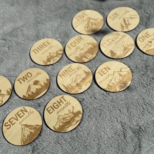 May include: A set of twelve wooden discs with engraved mountain and nature scenes. Each disc is labeled with a number and the word "months" or "year", from "One month" to "Eleven months" and "One year". The discs are arranged on a gray surface.