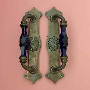 May include: Two matching door handles featuring a vertical brass backplate with a weathered patina and a central floral rosette. Each handle includes a faceted, deep blue glass grip section secured by brass hardware at the top and bottom ends.