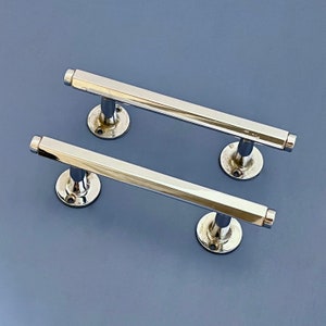 Può includere: Due maniglie per porte cromate lucide con un design a barra esagonale. Ogni maniglia ha una base circolare con tre fori per le viti. Le maniglie sono poste su uno sfondo blu-grigio.