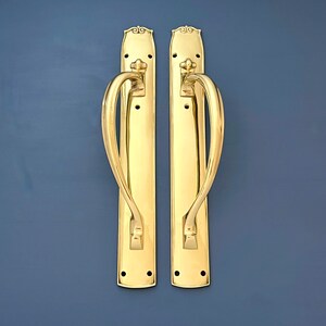 May include: Two gold door handles with ornate designs. The handles have a curved shape and are attached to rectangular plates.