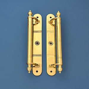 Könnte beinhalten: Zwei goldfarbene Türbänder mit dekorativen Details. Jedes Band hat eine abgerundete Ober- und Unterseite, mit einem leicht breiteren Mittelteil. Die Bänder sind an einer flachen, rechteckigen Platte befestigt.