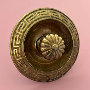 May include: Antique brass door knob with a decorative floral center and a Greek key border. The knob has a warm, aged patina, suggesting it is vintage. The design is symmetrical and detailed, suitable for vintage home decor.