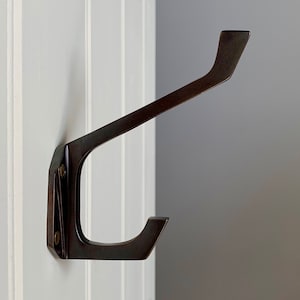 Puede incluir: Un gancho de metal negro con un diseño único. El gancho está montado en una pared blanca.