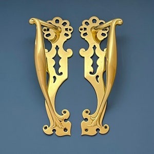 Puede incluir: Un par de tiradores de puerta de latón ornamentados con un diseño decorativo e intrincado. Los tiradores están hechos de latón macizo y tienen un acabado pulido.