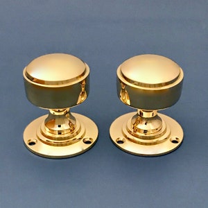 Può includere: Due pomelli per porte in ottone lucido con una parte superiore rotonda leggermente bombata e una base circolare. I pomelli hanno un anello rialzato attorno alla parte superiore.