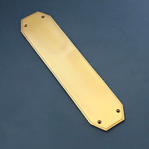 Puede incluir: Una placa de puerta de color dorado con forma rectangular y bordes redondeados. La placa tiene una superficie lisa y pulida y cuatro pequeños agujeros.