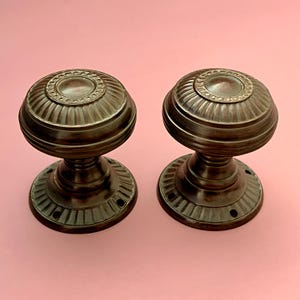 Pode incluir: Um par de maçanetas redondas de metal em tom de bronze escuro, com um design canelado e estriado na pega esférica e uma placa de base circular a condizer com orifícios para parafusos visíveis. O acabamento apresenta uma pátina metálica.