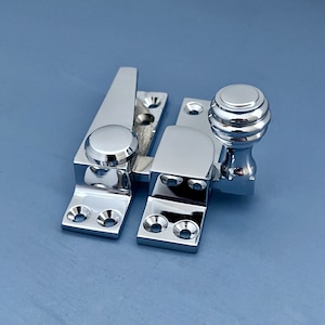 Könnte beinhalten: Ein polierter Fensterverschluss aus Chrom mit einem runden Knopf und einem Hebel. Der Verschluss hat eine glänzende, reflektierende Oberfläche und dient zur Sicherung eines Fensters. Die Hardware ist auf blauem Hintergrund montiert.