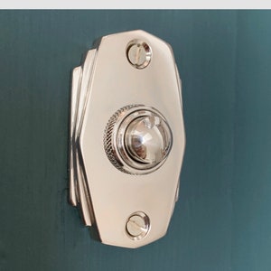 May include: A polished nickel door button with a rounded, slightly raised button in the center. The button is mounted on a rectangular plate with a geometric design.