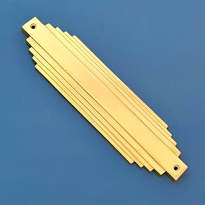 May include: A gold-coloured metal door plate with an Art Deco design. The plate has a rectangular shape with a series of horizontal lines that create a stepped effect.