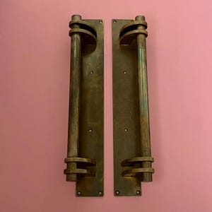 May include: Two antique brass door pulls with a vertical bar design and decorative circular accents. The pulls are mounted on rectangular backplates with visible screw holes. The background is a solid pink color.