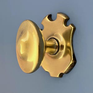 Könnte beinhalten: Nahaufnahme eines Türknaufs aus Messing mit einer schildförmigen Rückplatte. Der Knauf ist oval mit einem sternförmigen Design. Die Rückplatte hat einen dekorativen, gewellten Rand. Die Hardware ist goldfarben glänzend.