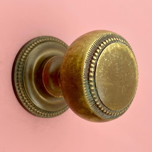 Könnte beinhalten: Nahaufnahme eines antiken Türknaufs aus Messing mit rundem, strukturiertem Design. Der Knauf hat eine kreisförmige Rückplatte mit einem Perlenrand. Das Metall hat eine warme, gealterte Patina, vor rosa Hintergrund.